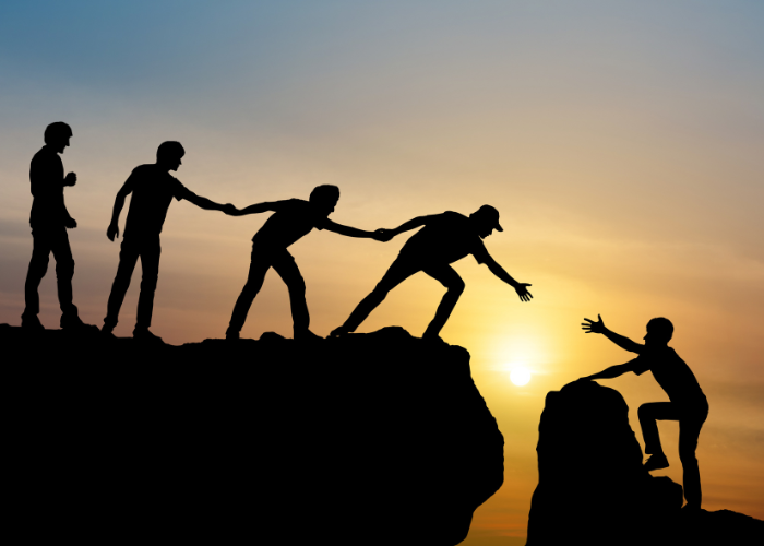 Silhouettes of four people on a cliff, holding hands to help another climb up, with a sunset in the background, symbolizing teamwork and support.